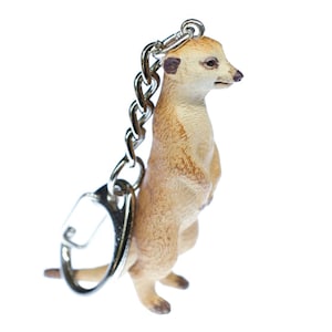 Puede incluir: Un llavero de plástico marrón y beige con forma de suricata realista. El suricata está de pie con las patas juntas y la cabeza ligeramente inclinada hacia un lado. El llavero está unido a una cadena y un anillo de metal plateado.