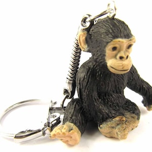 May include: A small, black and brown chimpanzee keychain. The chimpanzee is sitting with its legs crossed and its arms resting on its knees. The keychain has a silver spring clip and a silver ring.