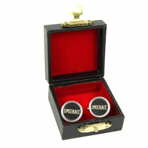 May include: A black cufflink box with a red velvet interior. The cufflinks are silver with black centres and the word "UMSCHALT" in gold lettering.