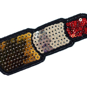 May include: A sequined patch in the shape of a lipstick. The patch is black with sections of gold, silver, and red sequins. The sequins are round and closely spaced, creating a shimmering effect. The patch is likely for clothing or accessories.