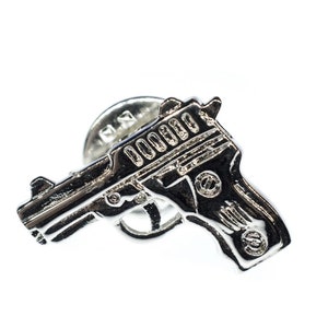 May include: A silver-coloured pistol-shaped pin with detailed features. The pin is set against a white background, emphasising its metallic shine and design. The pin is a small accessory.