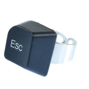 Könnte beinhalten: Ein schwarzer Plastikring mit einem silbernen Band. Der Ring ist in Form einer Computertaste geformt und trägt die Aufschrift "Esc".