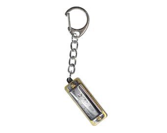 May include: A miniature harmonica keychain featuring a silver-toned metal cover plate with the text "SWAN" and "HARMONICA" engraved on the front. The instrument is attached to a silver-colored metal chain and a circular split ring key holder.