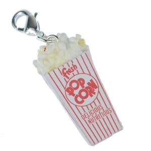 Könnte beinhalten: Ein silberner Anhänger in Form eines Popcorn-Bechers. Der Becher ist rot und weiß mit den Worten "Fresh Pop Corn" und "Delicious Nutritious" bedruckt. Der Anhänger hat einen silbernen Verschluss.