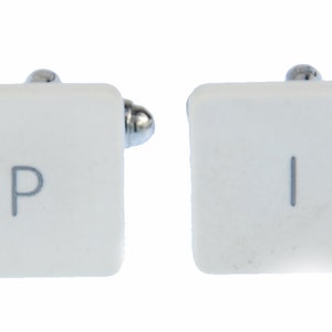 May include: A pair of white cufflinks with silver accents. The cufflinks are shaped like computer keys, one with the letter "P" and the other with the letter "I".