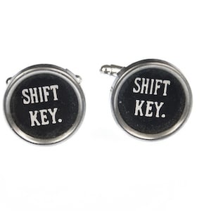 May include: A pair of silver cufflinks with black round buttons that say "SHIFT KEY." in white lettering.