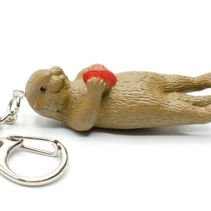 May include: A brown sea otter keyring holding a red heart. The keyring has a silver metal ring and chain.