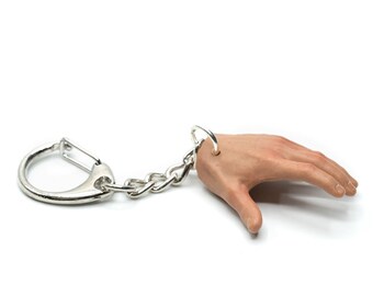 May include: A novelty keychain featuring a realistic, flesh-toned hand with a silver chain and clasp. The hand is in a relaxed position, with the thumb and index finger slightly apart. The keychain is designed to be attached to keys or bags.