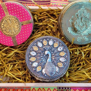 May include: Three round metal containers in a white tray filled with gold paper shreds. One is bright pink with a gold floral centre, one is teal with an embossed Ganesha figure, and one is silver with a detailed peacock design on the lid.