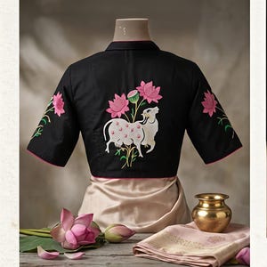 May include: A black short-sleeved blouse featuring intricate embroidery of a white cow and pink lotus flowers on the back. The garment has a collar and pink trim along the sleeve edges. The design includes green stems and leaves.