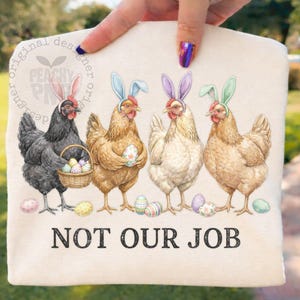 May include: A cream-colored tote bag featuring four chickens wearing bunny ears and holding Easter eggs. The text "NOT OUR JOB" is printed in black below the chickens. The bag has a whimsical, colorful design.
