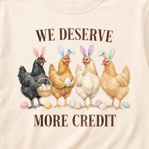 May include: Cream-colored t-shirt featuring a watercolor illustration of four chickens wearing Easter bunny ears. The chickens are surrounded by Easter eggs and the text "WE DESERVE MORE CREDIT" is printed above and below the image.