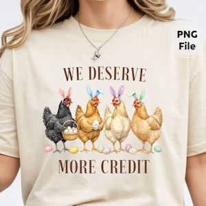 May include: Beige t-shirt featuring a watercolor illustration of four chickens wearing Easter bunny ears. The text "WE DESERVE MORE CREDIT" is printed above and below the chickens. Easter eggs are scattered around the chickens.
