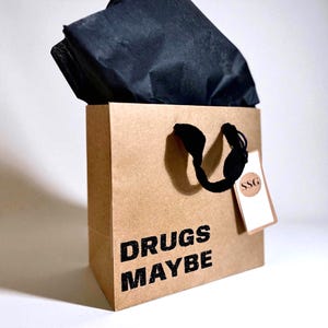 May include: A brown paper gift bag with black handles and the words "DRUGS MAYBE" printed in black. The bag contains black tissue paper and a tag with the letters "SSG".