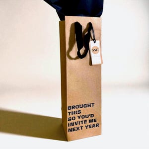 Könnte beinhalten: Eine braune Weintasche aus Papier mit schwarzen Griffen und dem Text "BROUGHT THIS SO YOU'D INVITE ME NEXT YEAR" in Schwarz gedruckt. Ein kleines Etikett mit den Buchstaben "SSG" hängt am Griff.