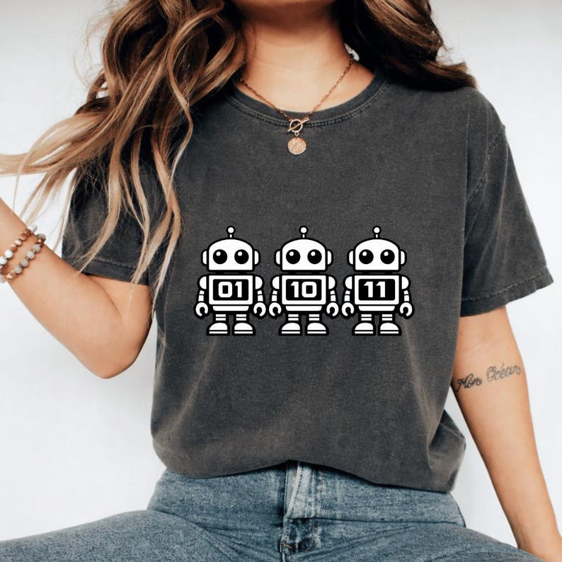 K&ouml;nnte beinhalten: Dunkelgraues T-Shirt mit drei wei&szlig;en Roboter-Illustrationen. Jeder Roboter hat eine andere Nummer auf der Brust: 01, 10 und 11. Das T-Shirt hat einen l&auml;ssigen Stil.