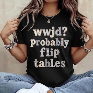 May include: Black t-shirt with the text "wwjd? Probably flip tables" in a mix of patterned and solid fonts. The shirt is a crew neck and appears to be made of a soft material.