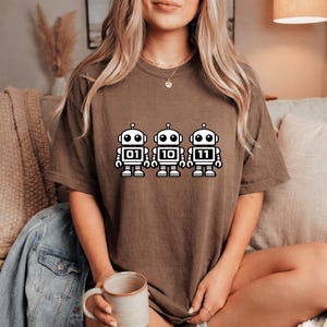 K&ouml;nnte beinhalten: Ein braunes T-Shirt mit drei wei&szlig;en Cartoon-Robotern, die die Bin&auml;rzahlen 01, 10 und 11 anzeigen. Die Roboter haben schwarze Umrisse und Details. Das T-Shirt ist einfarbig und hat eine lockere Passform.