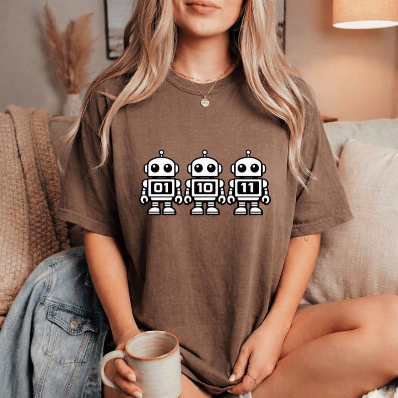 K&ouml;nnte beinhalten: Ein braunes T-Shirt mit drei wei&szlig;en Cartoon-Robotern, die die Bin&auml;rzahlen 01, 10 und 11 anzeigen. Die Roboter haben schwarze Umrisse und Details. Das T-Shirt ist einfarbig und hat eine lockere Passform.