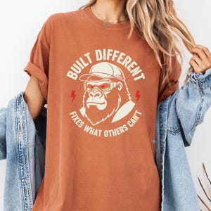 以下が含まれることがあります： キャップとゴーグルをつけたゴリラのグラフィックがプリントされた、錆色のTシャツ。「BUILT DIFFERENT FIXES WHAT OTHERS CAN'T」の文字。下部には「DIGITAL PRODUCT PNG」の文字。