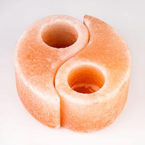 May include: A dual-piece candle holder crafted from pink Himalayan salt, shaped into a yin-yang symbol. The two interlocking curved sections each feature a circular opening at the top, designed to hold a candle within the textured, crystalline surface.