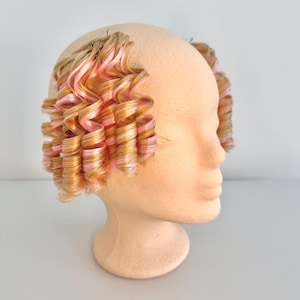 Puede incluir: Una cabeza de maniquí beige con dos secciones laterales de extensiones de cabello. El cabello está peinado en rizos en espiral apretados y presenta una mezcla de mechones rubio claro y rosa suave a lo largo de toda la longitud de los rizos.