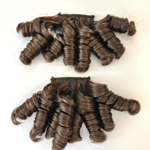 May include: Two sets of brown, tightly curled hair extensions. Each set features multiple ringlets attached to a black weft. The hair has highlights and lowlights, creating a dimensional look. The curls are uniform and well-defined.