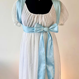 May include: A white dress with a swiss dot texture, featuring short puff sleeves, a scoop neckline, and a light blue satin sash tied in a large bow at the waist. The sash has long, flowing ends finished with small, light brown decorative tassels.