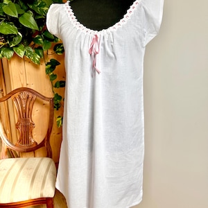 May include: A white knee-length nightgown featuring short sleeves and a scoop neckline. The neckline is detailed with white eyelet lace trim woven with a thin pink ribbon that ties into a small bow at the center front of the garment.