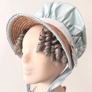 Könnte beinhalten: Hellblaue Haube mit Strohschirm und grauen Locken. Die Haube hat ein weißes Futter und ein hellblaues Band. Die Haube befindet sich auf einem Schaufensterkopf.