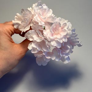 Könnte beinhalten: Ein Bund zarter, hellrosa Kunstblumen mit mehrfach geschichteten Blütenblättern. Die Blumen werden in einer Hand vor einem schlichten grauen Hintergrund gehalten, der einen Schatten wirft. Die Blüten haben ein weiches, romantisches Aussehen, geeignet für Blumengestecke oder Bastelprojekte.