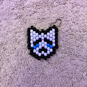 May include: A beaded cat-shaped keychain. The cat design features black, white, and blue beads. The keychain has a silver-colored metal clasp for attachment.