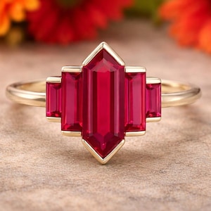 May include: A gold ring featuring a geometric design with a central, elongated, rectangular ruby gemstone, flanked by two smaller rubies on each side. The rubies are a vibrant red color, set against a gold band.