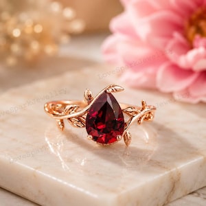 Può includere: Un anello in oro rosa con una pietra preziosa rosso scuro a forma di pera come pietra centrale. La fascia è decorata con intricati motivi a foglia in metallo che si estendono dall'incastonatura lungo entrambi i lati dell'anello.
