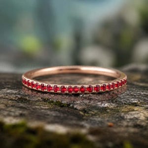 Könnte beinhalten: Ein roségoldener Ring mit einer durchgehenden Reihe kleiner, leuchtend roter Edelsteine. Der Ring ist auf einem strukturierten, natürlichen Hintergrund platziert, der das Funkeln des Schmucks hervorhebt. Die Edelsteine sind gleichmäßig angeordnet.