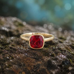 Pode incluir: Um anel de ouro com uma grande pedra preciosa vermelha escura de corte quadrado. O anel está sobre uma superfície texturizada e musgosa, com um fundo desfocado verde e azul. A banda do anel é fina e polida.
