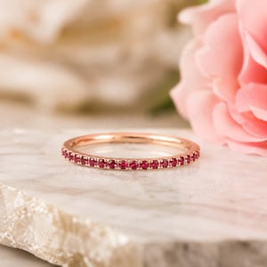 May include: A thin rose gold band ring featuring a continuous row of small, round, vibrant red rubies set in a shared prong setting. The ring is positioned horizontally, showcasing the uniform size and deep red color of the gemstones.