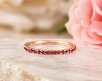 May include: A thin rose gold band ring featuring a continuous row of small, round, vibrant red rubies set in a shared prong setting. The ring is positioned horizontally, showcasing the uniform size and deep red color of the gemstones.