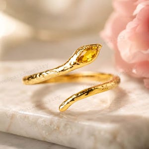 May include: A gold-toned open ring shaped like a snake with a textured, uneven surface. The head of the snake features a single marquise-cut yellow gemstone set into the metal. The ring has a bypass design that wraps around the finger.