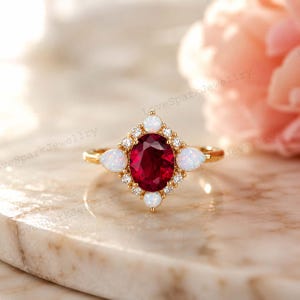 May include: A gold ring featuring a central oval-cut deep red gemstone surrounded by a halo of small round white diamonds and four pear-shaped white opals at the cardinal points, creating a geometric, star-like cluster design.