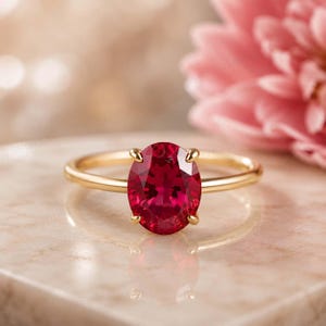Puede incluir: Un anillo de oro con una gran piedra preciosa de color rojo intenso y corte ovalado, sujeta por cuatro delicadas garras. La banda es fina y pulida, mostrando un perfil suave y redondeado que complementa la vibrante piedra central facetada.
