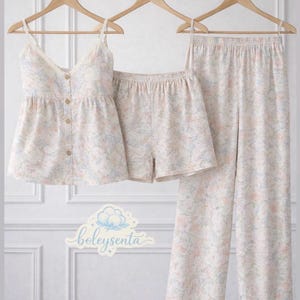 May include: A three-piece pajama set with a floral pattern in soft pastel colors. The set includes a camisole top with lace trim, button details, shorts, and long pants, all hanging on wooden hangers.