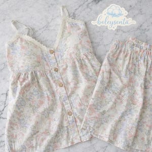 May include: A two-piece pajama set featuring a floral pattern in pastel colors. The top is a camisole style with lace trim and button closures. The matching shorts have an elastic waistband. The brand name "boleysenta" is visible in the upper right corner.