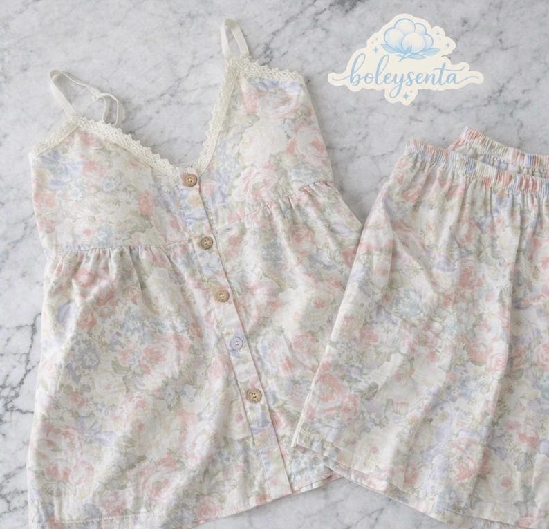 May include: A two-piece pajama set featuring a floral pattern in pastel colors. The top is a camisole style with lace trim and button closures. The matching shorts have an elastic waistband. The brand name "boleysenta" is visible in the upper right corner.