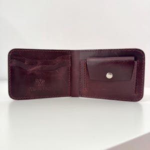 Puede incluir: Cartera de cuero burdeos abierta con múltiples ranuras para tarjetas y un monedero asegurado con un broche plateado. La cartera tiene el texto en relieve "Wrap & Fold".