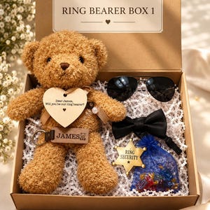 Puede incluir: Un oso de peluche marrón en una caja de cartón etiquetada RING BEARER BOX 1. El oso lleva una etiqueta de madera en forma de corazón que dice Dear James, Will you be our ring bearer? Incluye gafas de sol negras, una pajarita negra, un llavero con el nombre JAMES y una insignia de estrella dorada que dice RING SECURITY junto a una bolsa de organza azul.