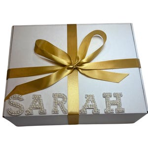 Op de afbeelding: Een witte geschenkdoos vastgebonden met een gouden satijnen lint en strik. De naam "SARAH" is gespeld in zilveren letters met parelachtige versieringen over de voorkant van de doos. De doos is gesloten.