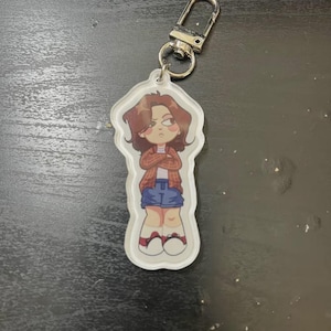 Puede incluir: Un llavero acrílico transparente con una ilustración de dibujos animados de una persona con cabello castaño, que lleva una camisa a cuadros roja, una falda vaquera azul y zapatillas blancas. El llavero tiene un cierre plateado.