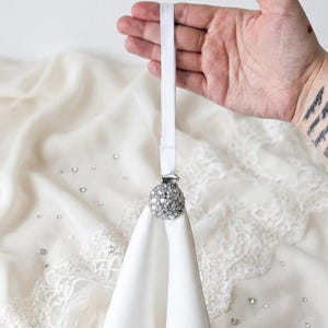 May include: A white fabric napkin held by a circular napkin ring featuring a cluster of clear, sparkling rhinestones. The ring is attached to a thin, white elastic band, designed to secure the folded fabric in a neat, elegant presentation.