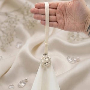 May include: A cream-colored fabric strap attached to a circular clip adorned with white faux pearls and clear crystals. The clip secures a piece of matching cream-colored fabric, creating a decorative accessory with a polished and elegant appearance.